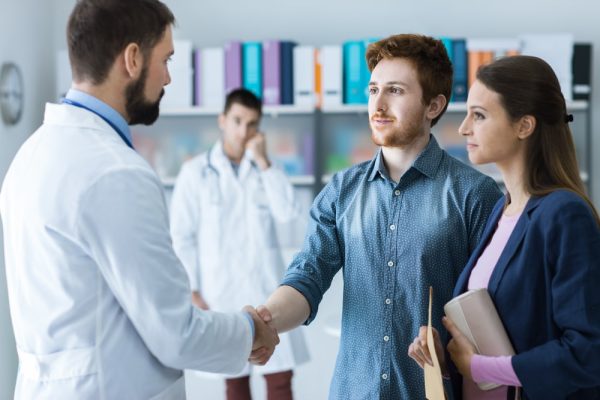 Casal heterossexual branco conversando com um médico branco, e outro médico branco ao fundo da imagem. O marido e o médico estão apertando as mãos.