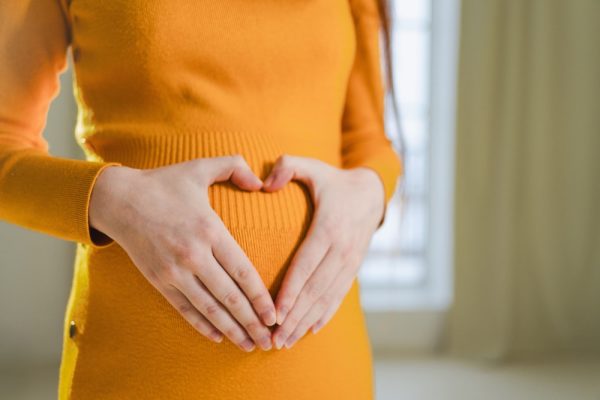 Mulher grávida com vestido laranja e mãos formando um coração na frente da barriga.