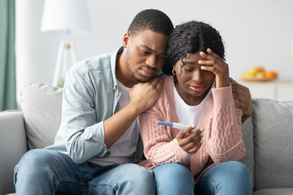 Casal negro e hétero. A mulher está segurando um exame de gravidez na mão e os dois estão olhando fixamente para o exame com um semblante de frustração.